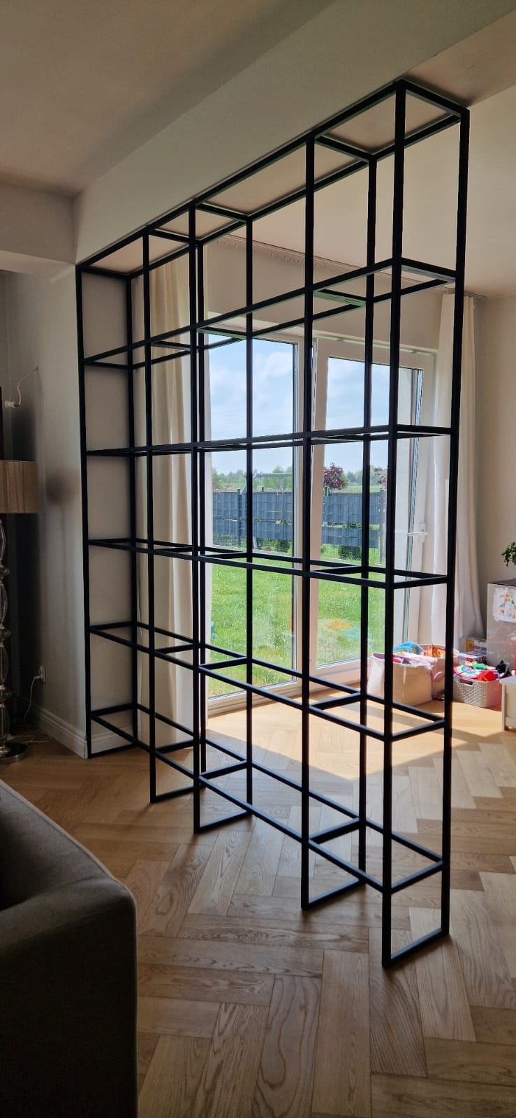 Metalowa konstrukcja w kształcie sześcianów, dzieląca przestrzeń w nowoczesnym wnętrzu z parkietem w jodełkę, widok na ogród przez okno balkonowe.
