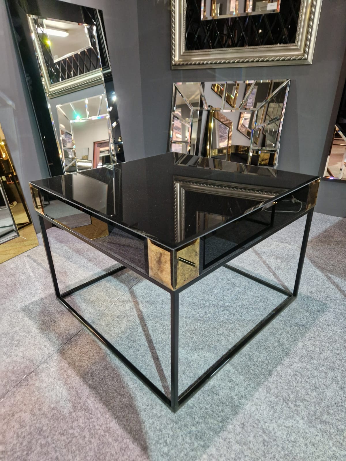 Stolik kawowy z czarnym, błyszczącym blatem i metalową ramą, z elementami dekoracyjnymi w kolorze złotym, na tle ściany z lustrami o różnych kształtach.