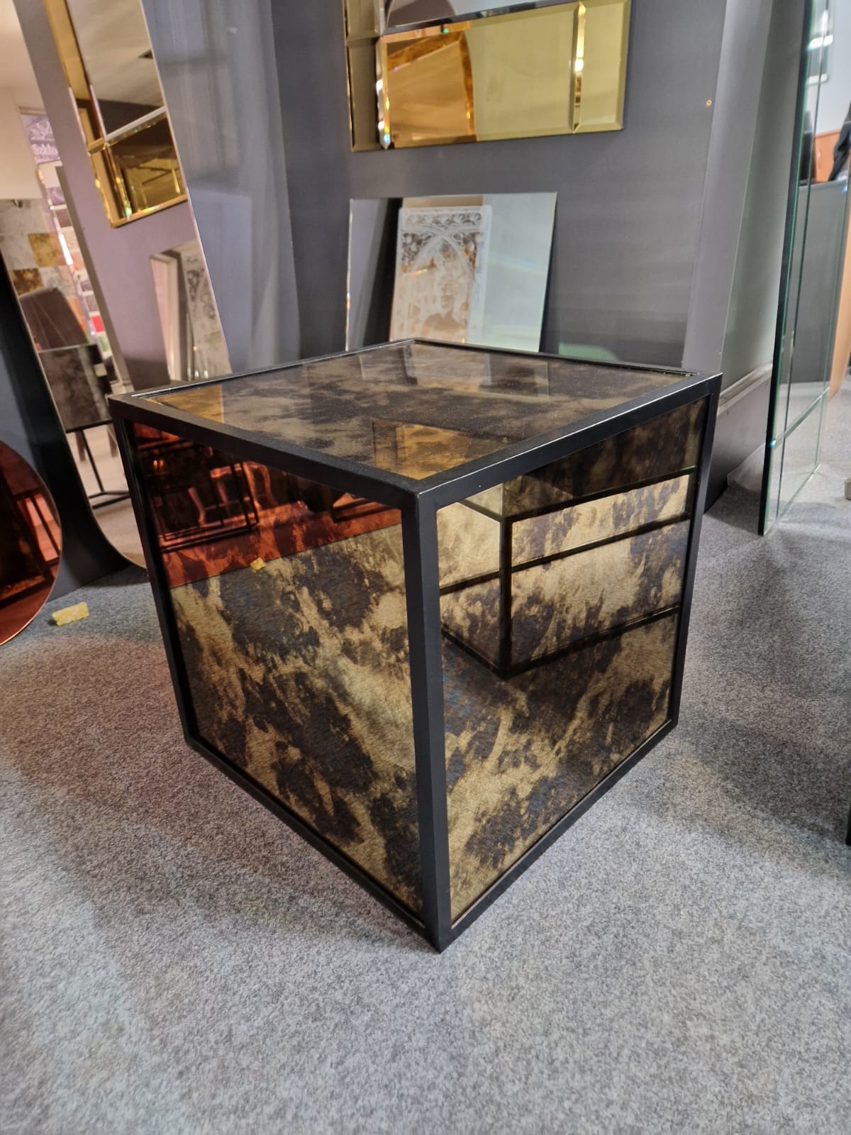 Kwadratowy stolik pomocniczy z czarną ramą i lustrzanymi ściankami o nieregularnym wzorze, stojący na szarym dywanie, z lustrami w tle.