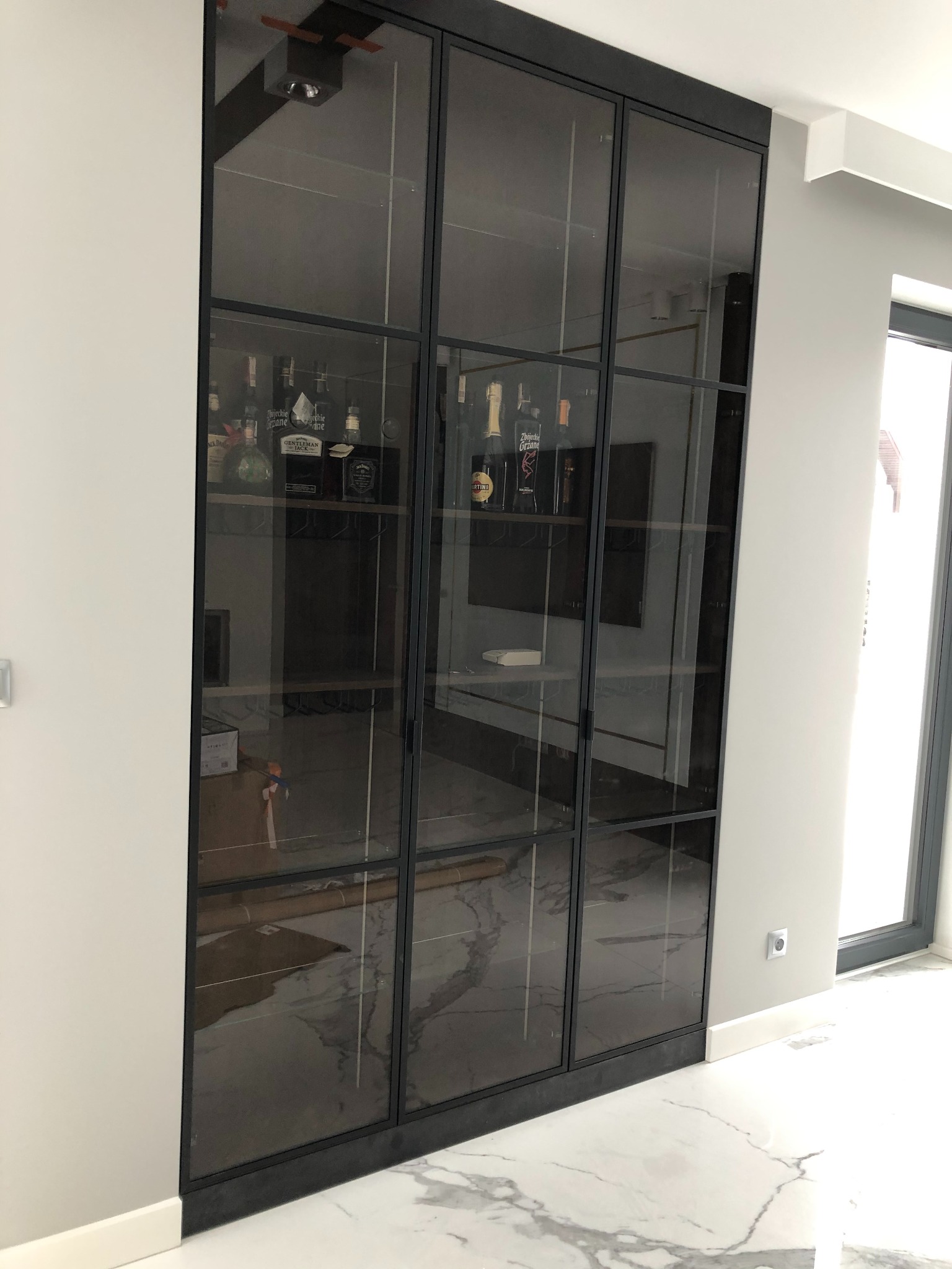 Nowoczesna, przeszklona witryna z czarnymi metalowymi ramami, prezentująca kolekcję alkoholi na marmurowej podłodze.