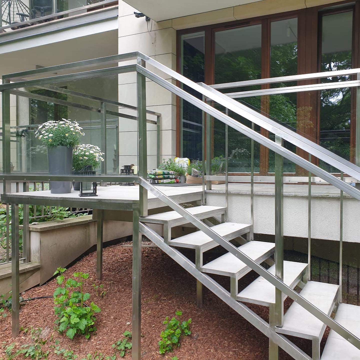Zewnętrzne schody metalowe z balustradą nierdzewną prowadzące na podest z kwiatami w donicach, widok z dołu na tle budynku.