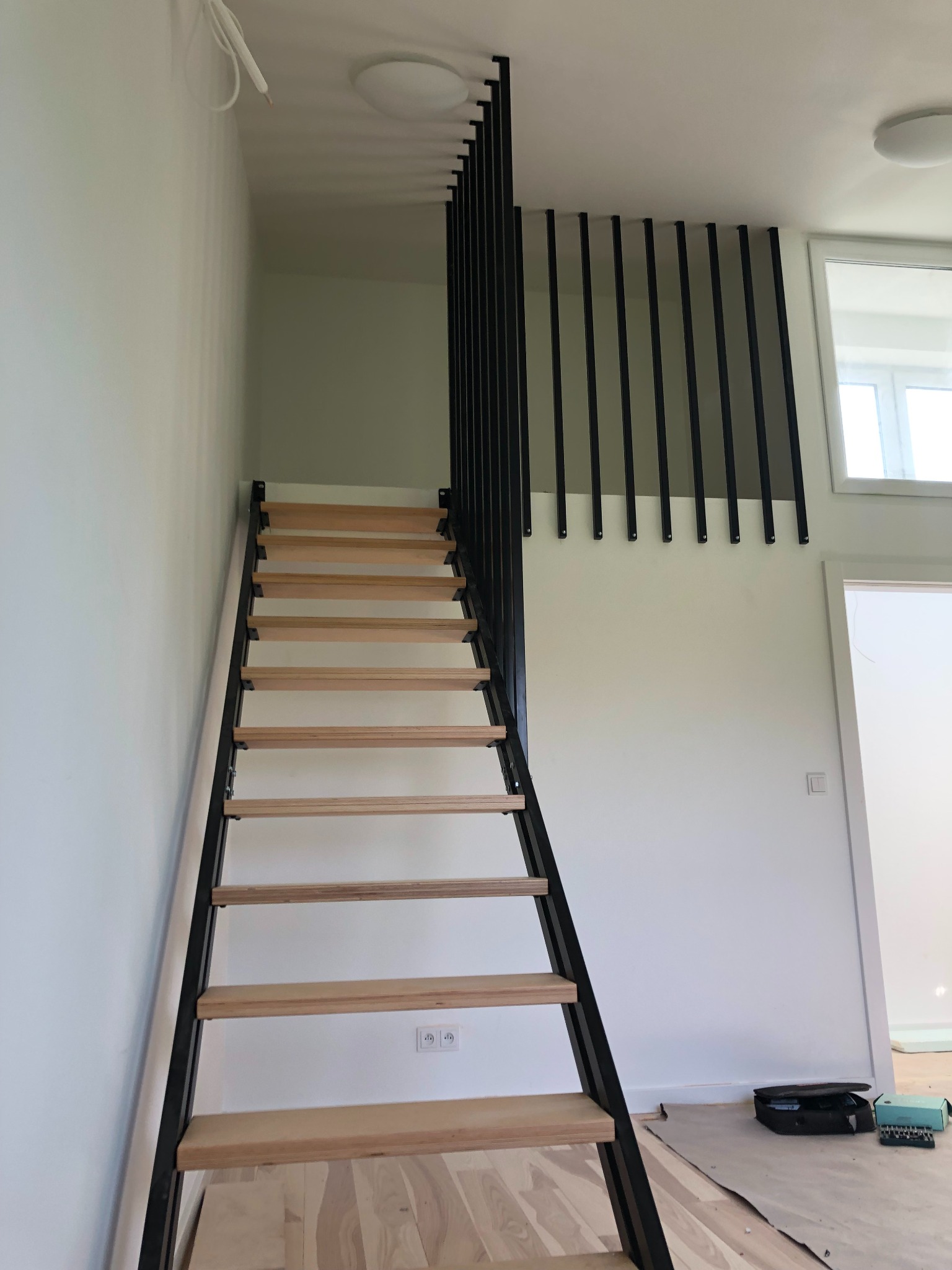 Nowoczesne schody z drewnianymi stopniami i czarną, metalową konstrukcją, widoczne z dołu, z minimalistyczną balustradą na antresoli i narzędziami na podłodze.