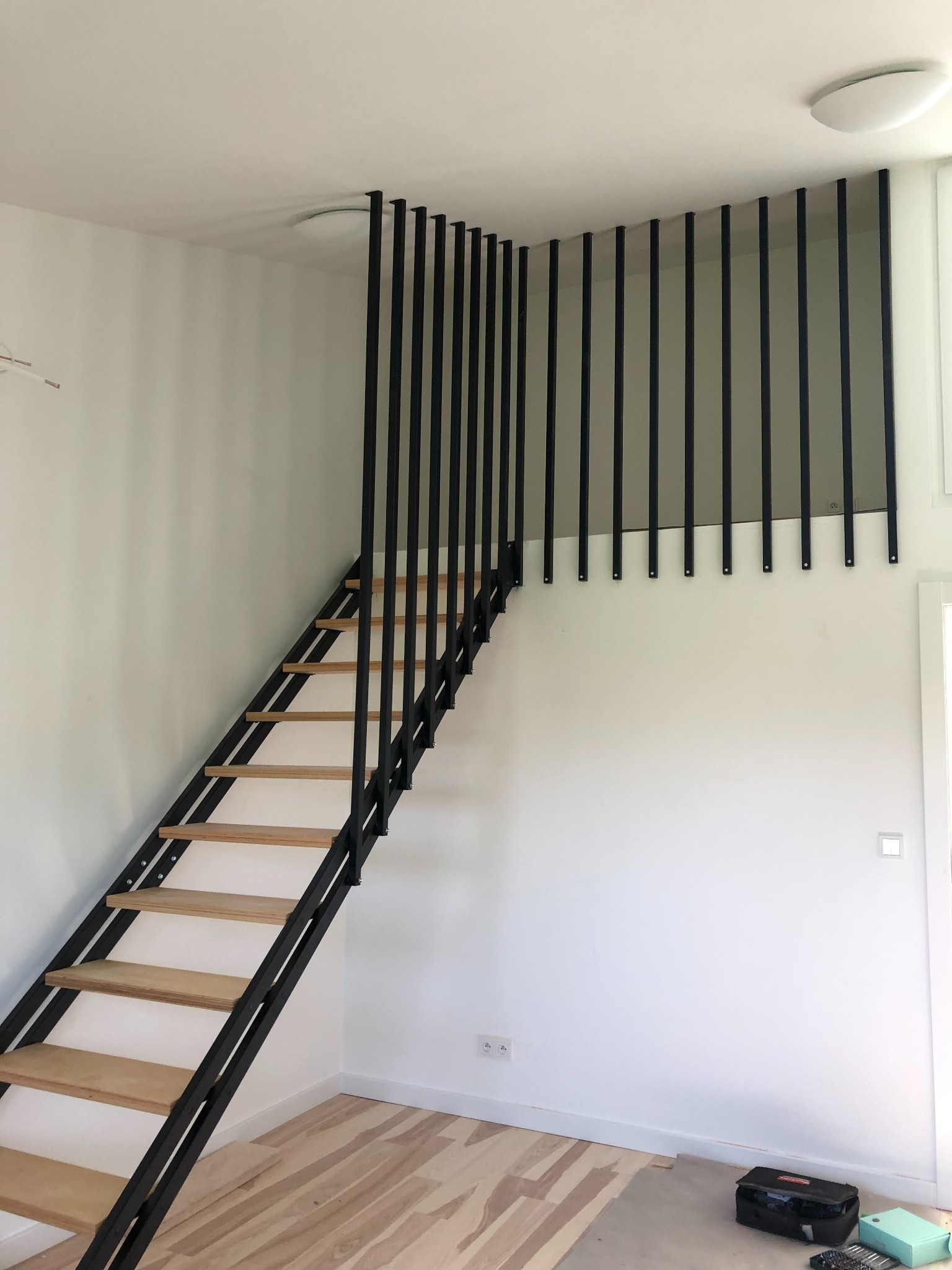 Metalowe schody z drewnianymi stopniami prowadzące na antresolę, zabezpieczone czarnymi, pionowymi prętami, widok z dołu, w tle białe ściany i podłoga w trakcie wykańczania.