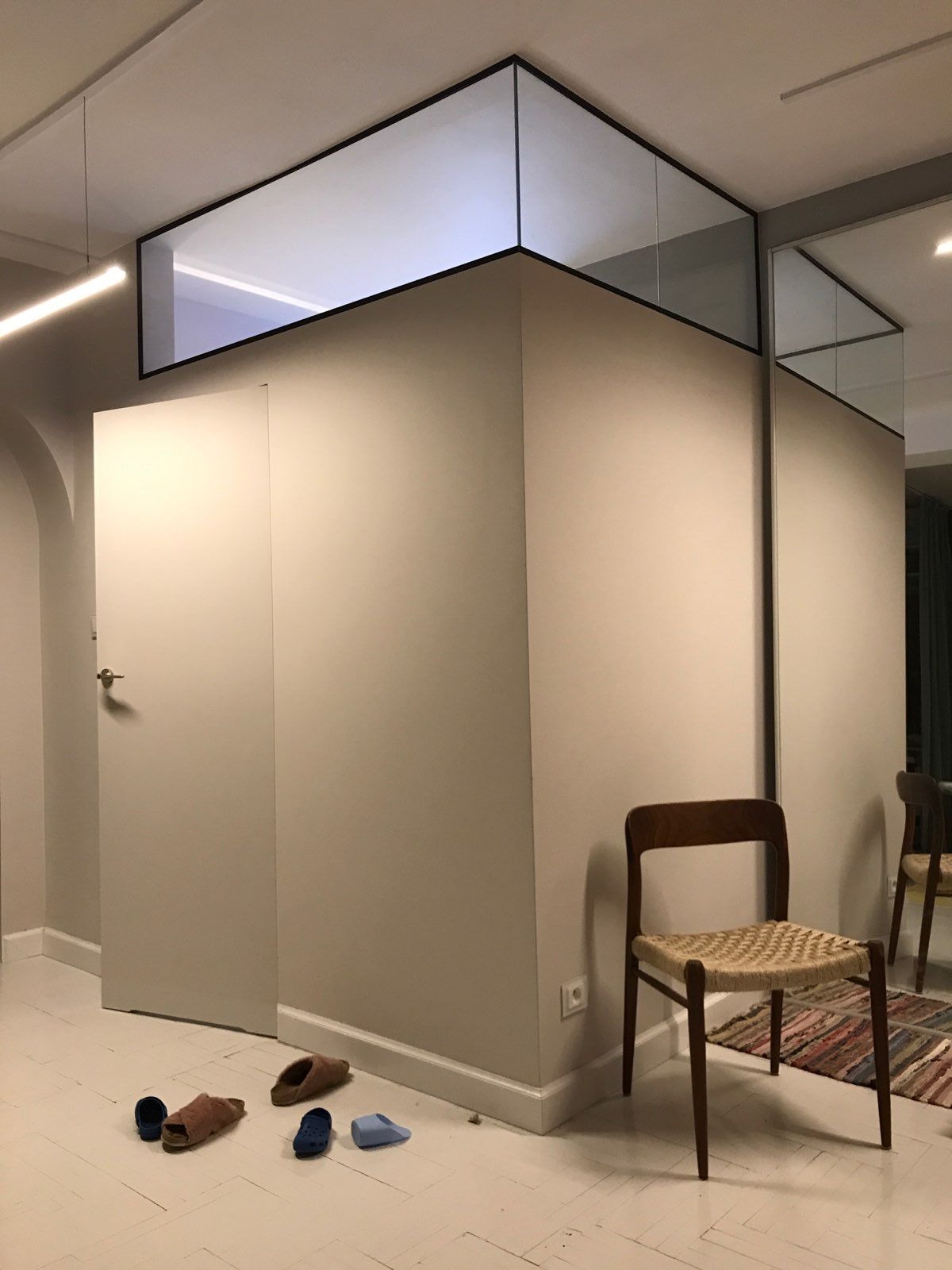 Nowoczesne wnętrze z zabudową antresoli nad ścianą, szklana konstrukcja z czarną ramą, drewniane krzesło z plecionym siedziskiem i porozrzucane kapcie na podłodze.