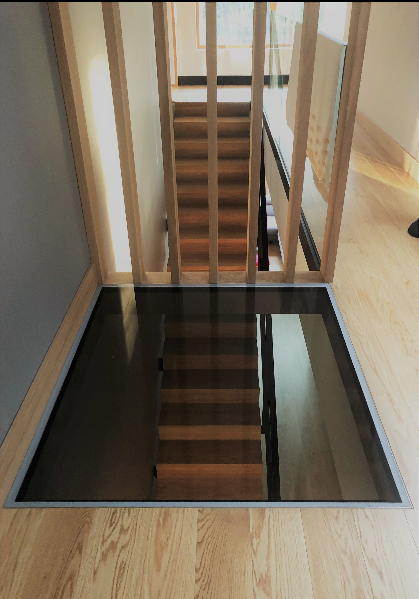Szklana podłoga nad schodami z drewnianą balustradą, widok z góry na drewniane stopnie odbijające się w tafli szkła.