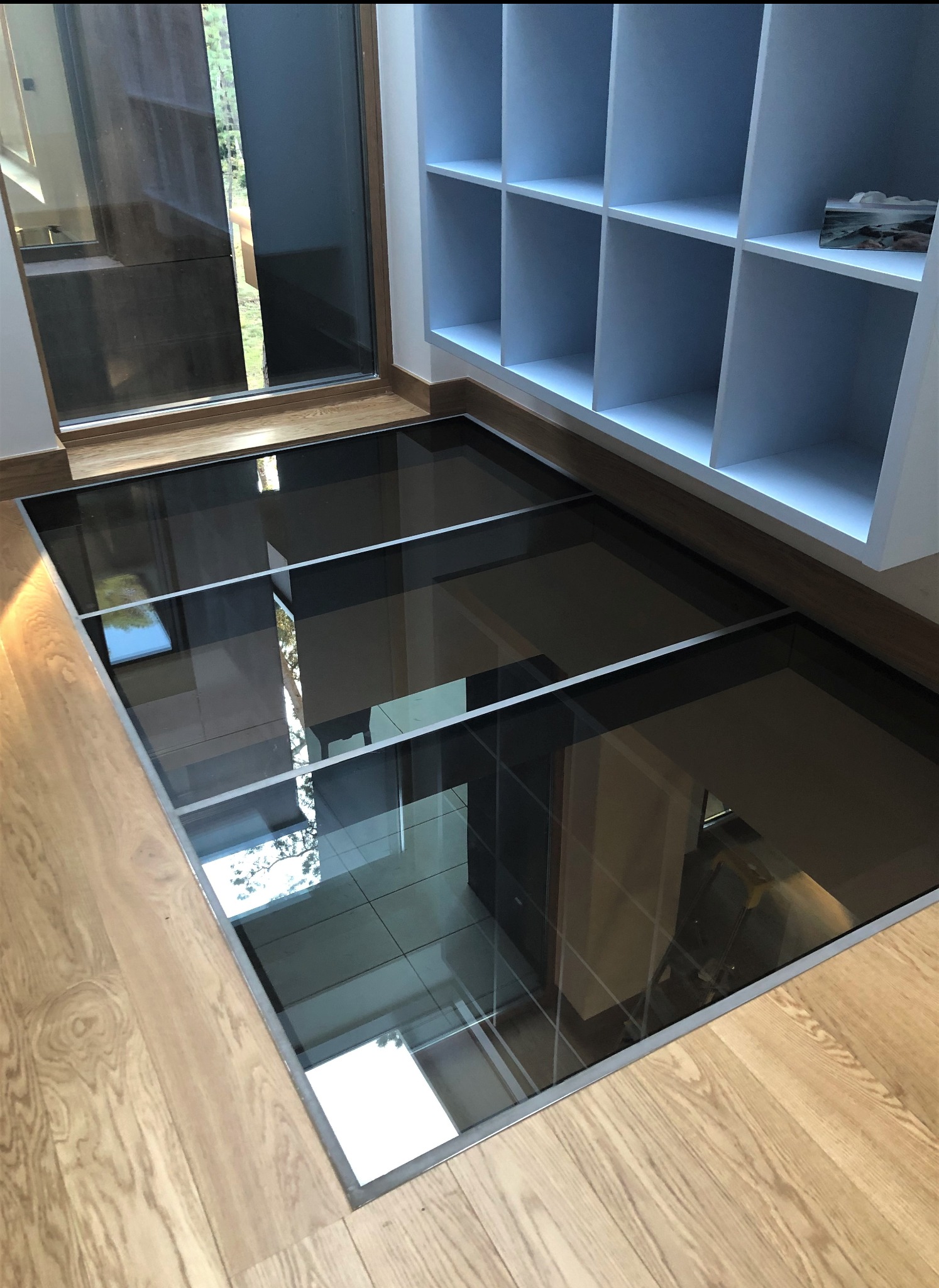 Szklana podłoga złożona z kilku paneli, wpuszczona w drewnianą podłogę, widok z góry, w tle białe półki.