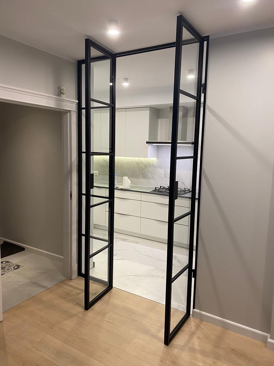 Dwuskrzydłowe, przeszklone drzwi z czarnymi, metalowymi szprosami oddzielające przedpokój z drewnianą podłogą od jasnej kuchni z marmurową podłogą i białymi meblami.