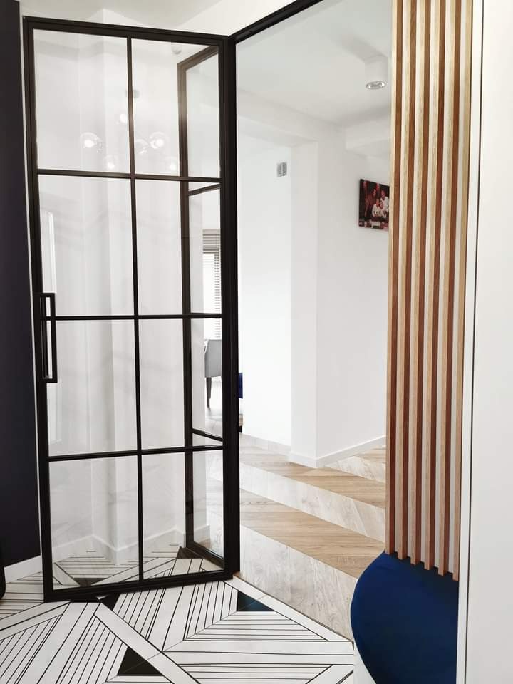 Szklana ściana działowa z czarnymi szprosami, otwierana jako drzwi, na tle wzorzystej podłogi i drewnianej okładziny ściennej.