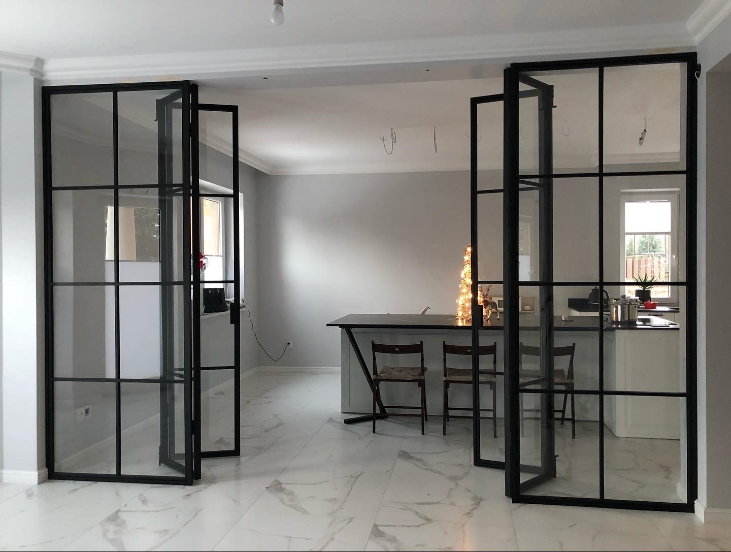 Nowoczesne, przeszklone drzwi wewnętrzne z czarnymi szprosami, dzielące przestrzeń salonu i kuchni, z widoczną podłogą z białego marmuru z szarymi żyłami i minimalistycznym wystrojem wnętrza.