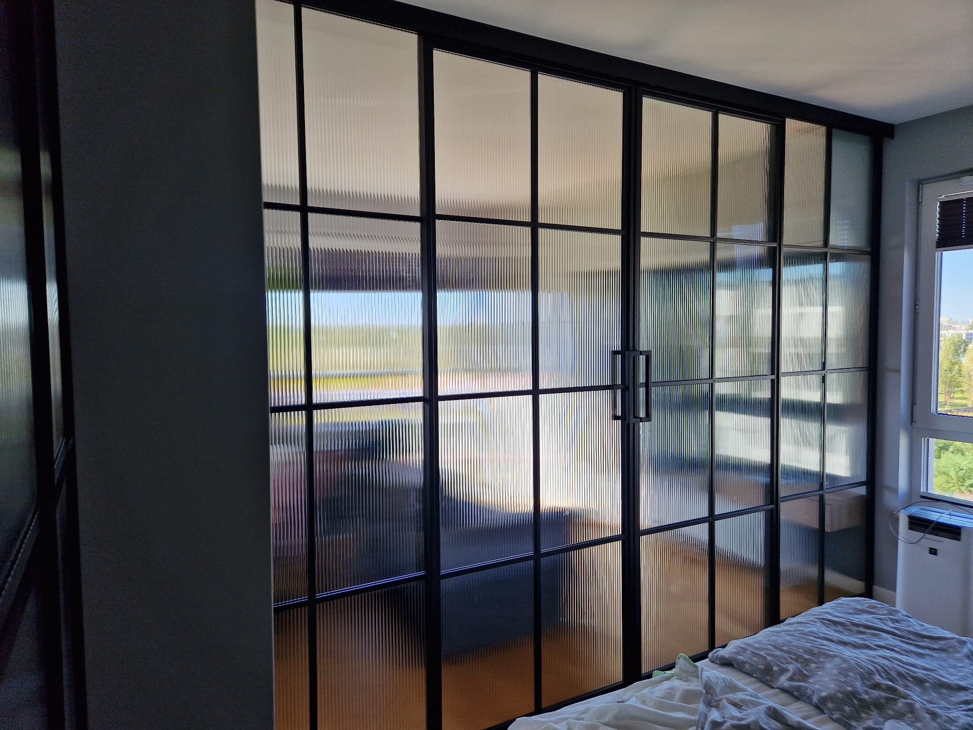 Przeszklona ściana działowa z czarnymi profilami i ryflowanym szkłem, oddzielająca sypialnię od innego pomieszczenia, widok na zewnątrz przez okno.