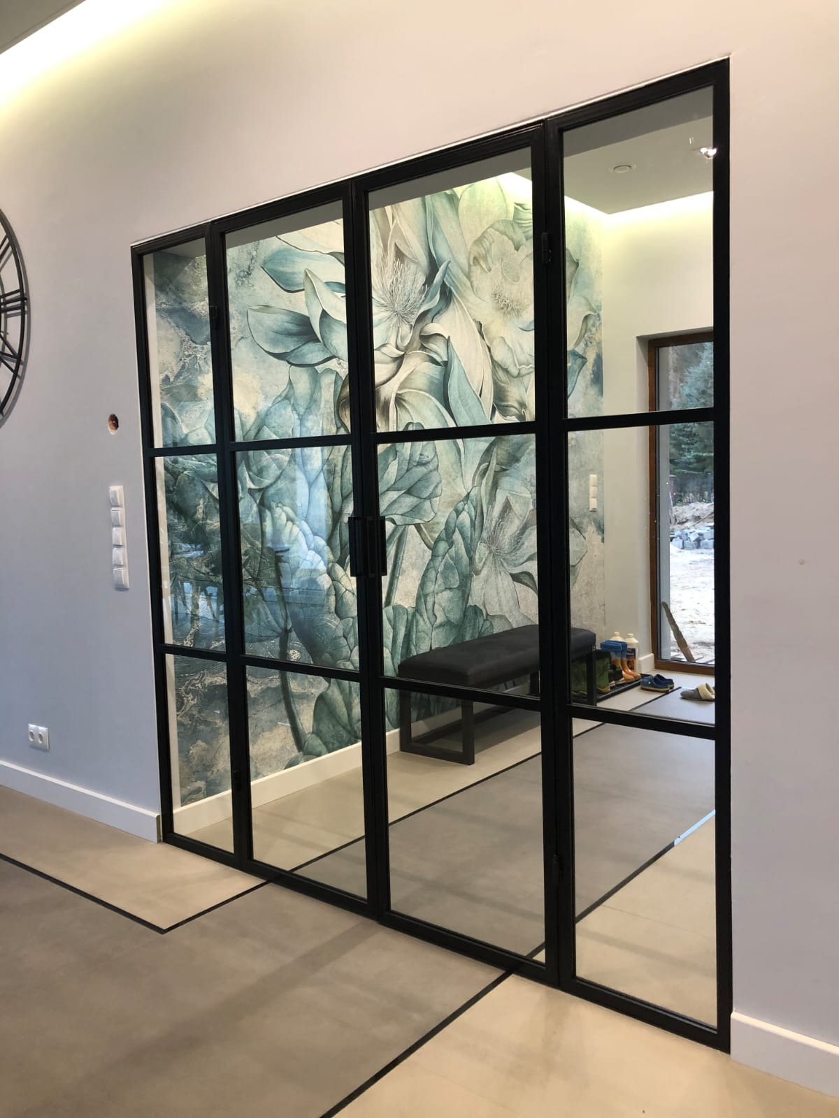 Nowoczesne, przeszklone drzwi wewnętrzne z czarnymi szprosami dzielącymi szkło na prostokątne pola, widok na korytarz z tapetą w kwiaty i ławką.