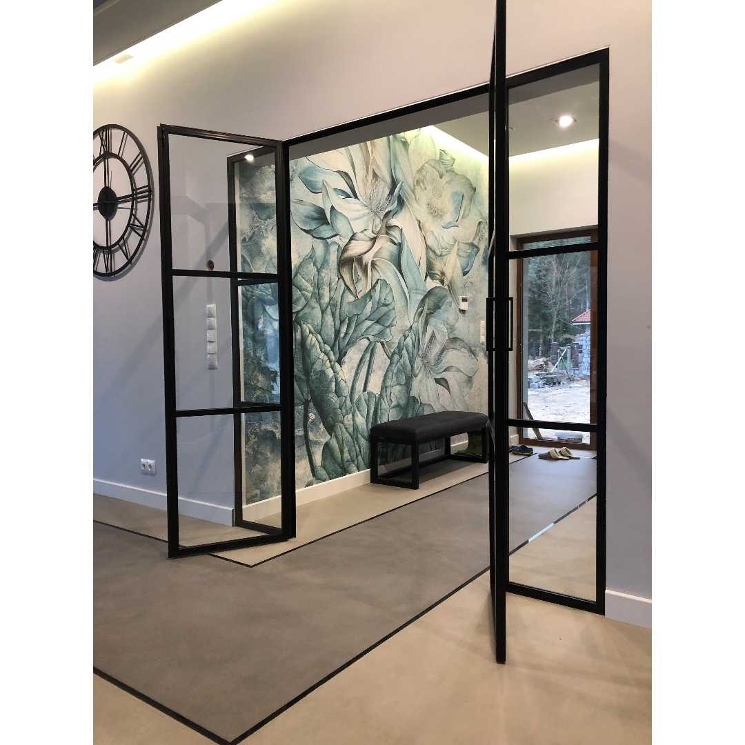 Nowoczesne, przeszklone drzwi wewnętrzne z czarnymi szprosami, otwarte na oścież, z widokiem na fototapetę z motywem kwiatowym i ławkę w tle, a także na zegar ścienny po lewej stronie.