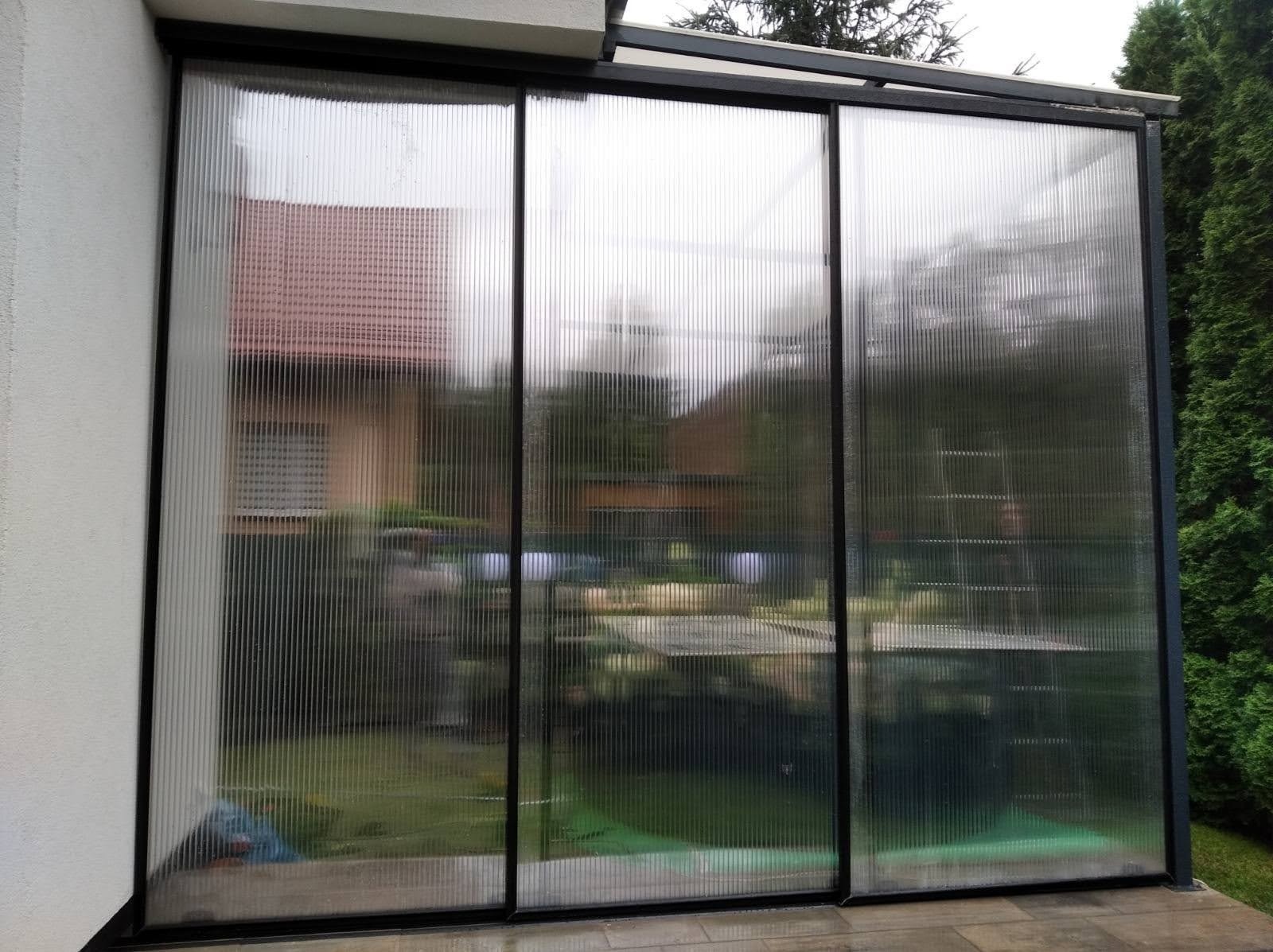 Zadaszenie tarasu z poliwęglanu z czarną metalową ramą, widok z zewnątrz na ogród i dom przez półprzezroczyste ściany.