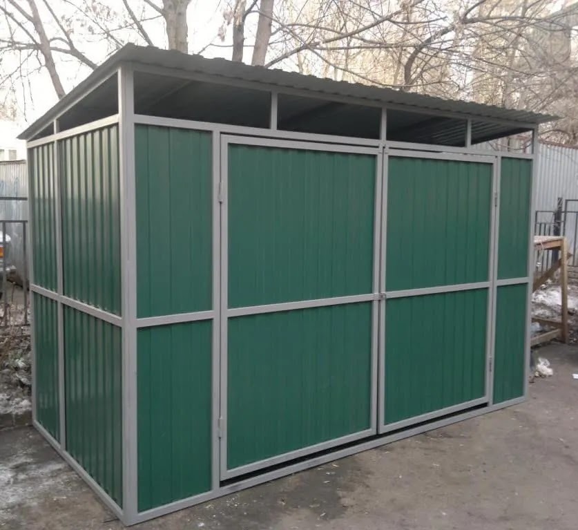 Zadaszona konstrukcja z zielonych paneli i szarej metalowej ramy, prawdopodobnie osłona na śmietniki, na tle drzew bez liści i fragmentu ogrodzenia.