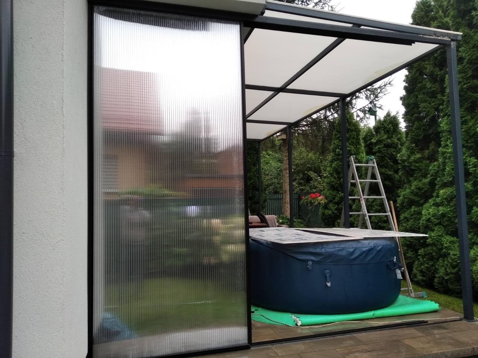 Zadaszona metalowa konstrukcja z przesuwną ścianą z poliwęglanu, pod spodem jacuzzi, w tle drabina oparta o konstrukcję.