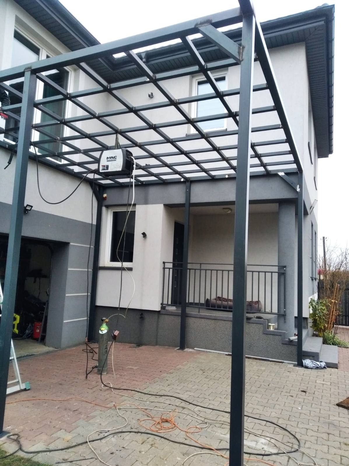 Metalowa pergola w trakcie budowy przy domu jednorodzinnym, widoczny spawacz i butla z gazem, rozłożone kable na kostce brukowej.