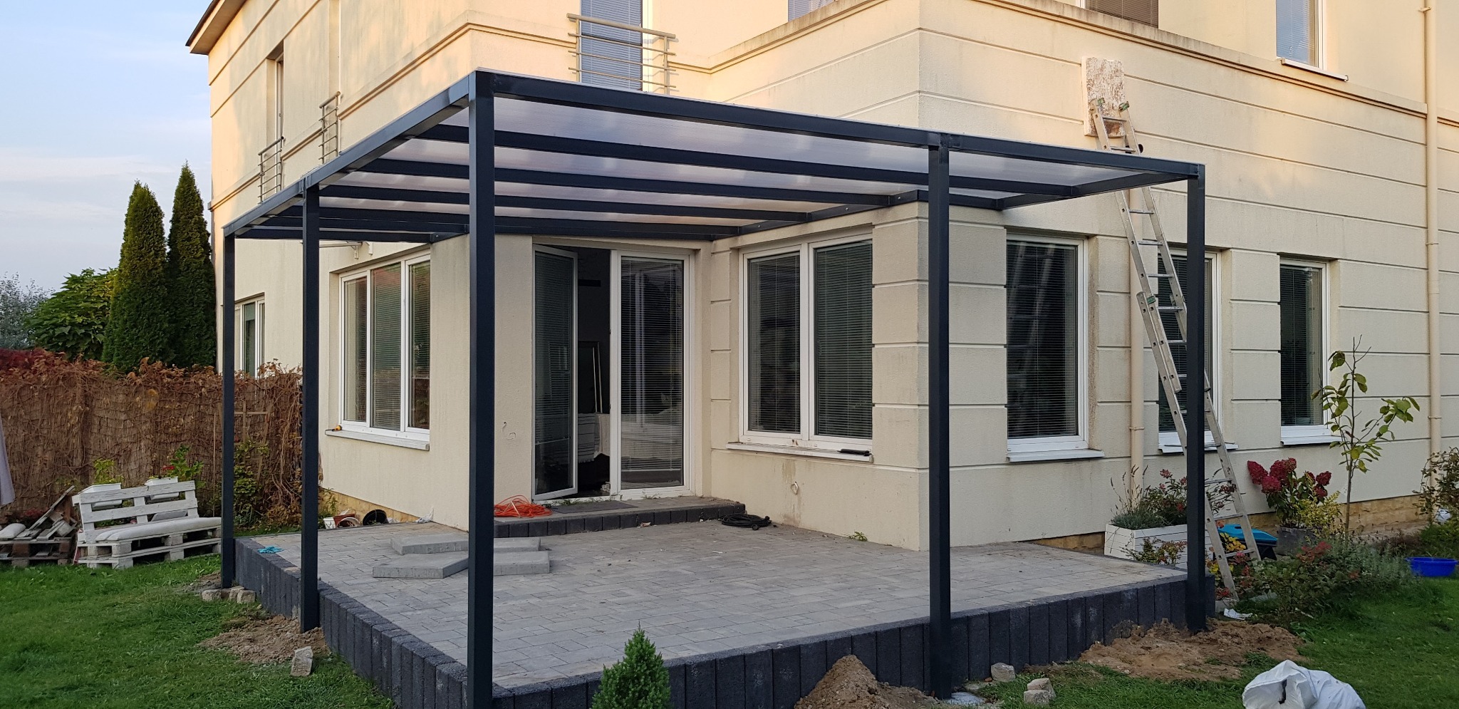 Nowoczesna, grafitowa pergola z przezroczystym zadaszeniem przylegająca do jasnego domu z oknami, widoczna drabina oparta o ścianę, świeżo ułożona kostka brukowa na tarasie.