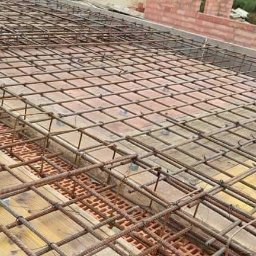 Grzegorz Grzegorz - Zbrojenie stropu ułożone na deskowaniu i pustakach szalunkowych, widoczne fragmenty muru z czerwonej cegły w tle.