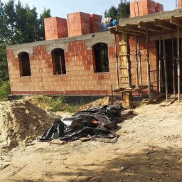 Grzegorz Grzegorz - Budowa domu w trakcie wznoszenia ścian z czerwonej cegły, widoczne otwory okienne z łukowym wykończeniem, na górze cegły w pomarańczowych opakowaniach, na ziemi czarna folia i piasek.