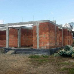 Grzegorz Grzegorz - Surowy stan budynku w trakcie wznoszenia, widoczne ściany z czerwonej cegły i betonowe słupy nośne, przygotowane materiały budowlane na zewnątrz, w tle zabudowania mieszkalne i drzewa.