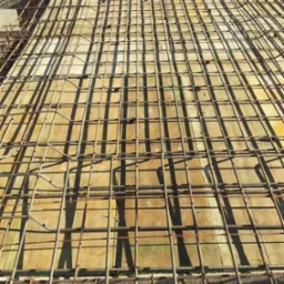 Zbrojenie fundamentów ułożone na żółtych szalunkach, widok z góry na siatkę z prętów stalowych, w tle trawa i budynek gospodarczy.