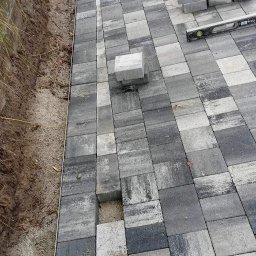 Grzegorz Grzegorz - Układanie szarej kostki brukowej na podsypce piaskowej, widoczna poziomica i stos kostki, krawężnik oddzielający od ziemi.