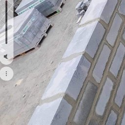 Grzegorz Grzegorz - Wznoszenie muru z szarych bloczków betonowych na fundamencie, widoczne palety z zapakowanymi bloczkami w tle