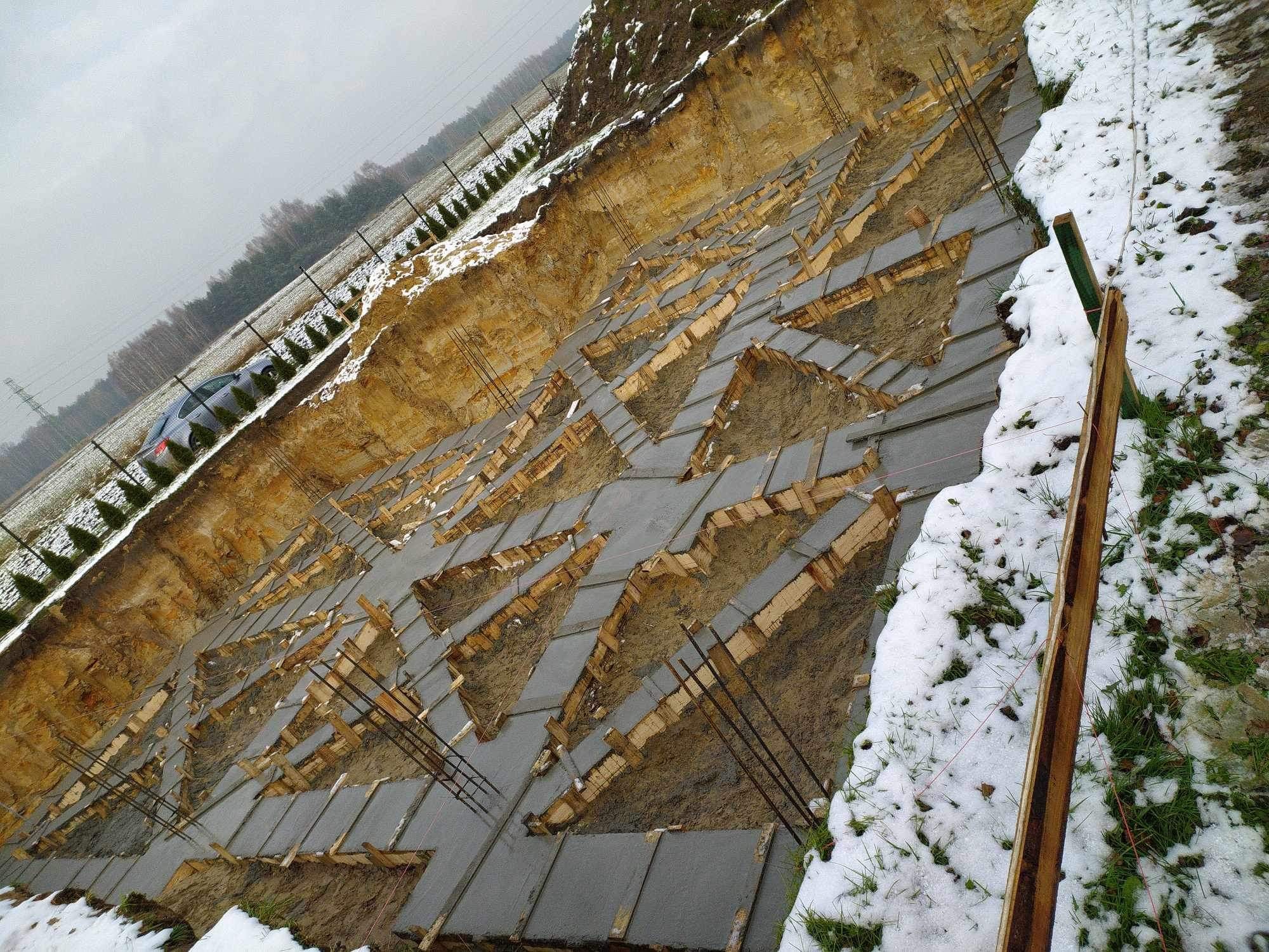 Przygotowanie fundamentów pod budynek w zimowej scenerii: wykop z drewnianym szalunkiem i zbrojeniem wypełniony betonem, częściowo przykryty śniegiem.