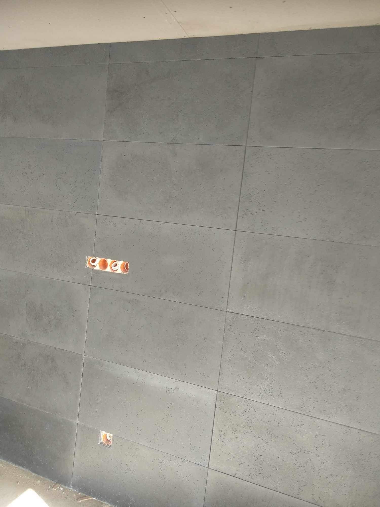 Ściana wyłożona szarymi, podłużnymi płytkami z widocznymi puszkami instalacyjnymi na różnej wysokości.