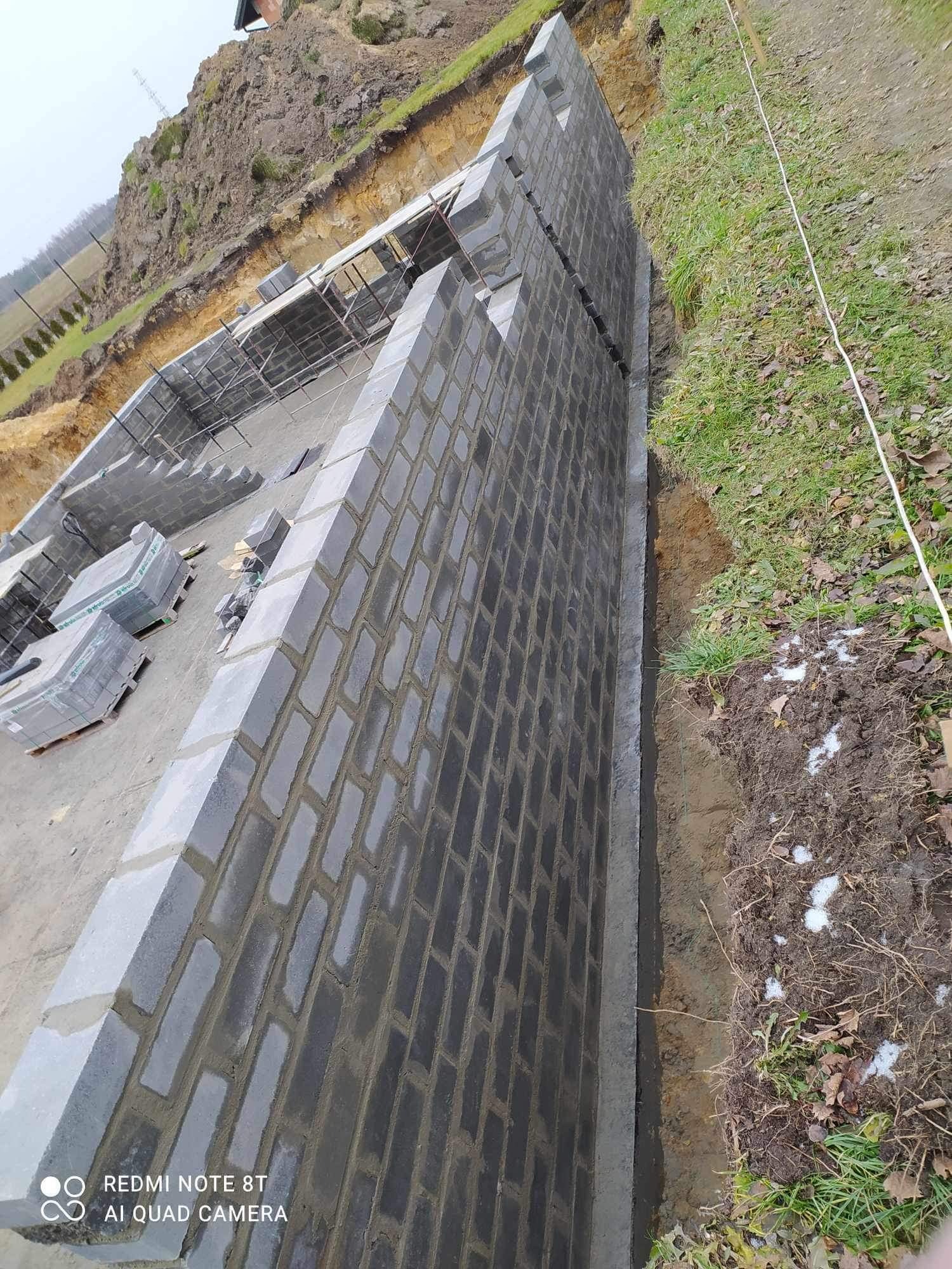 Murowanie ściany fundamentowej z bloczków betonowych, widoczne przygotowane podłoże i częściowo wzniesiona konstrukcja szkieletowa.