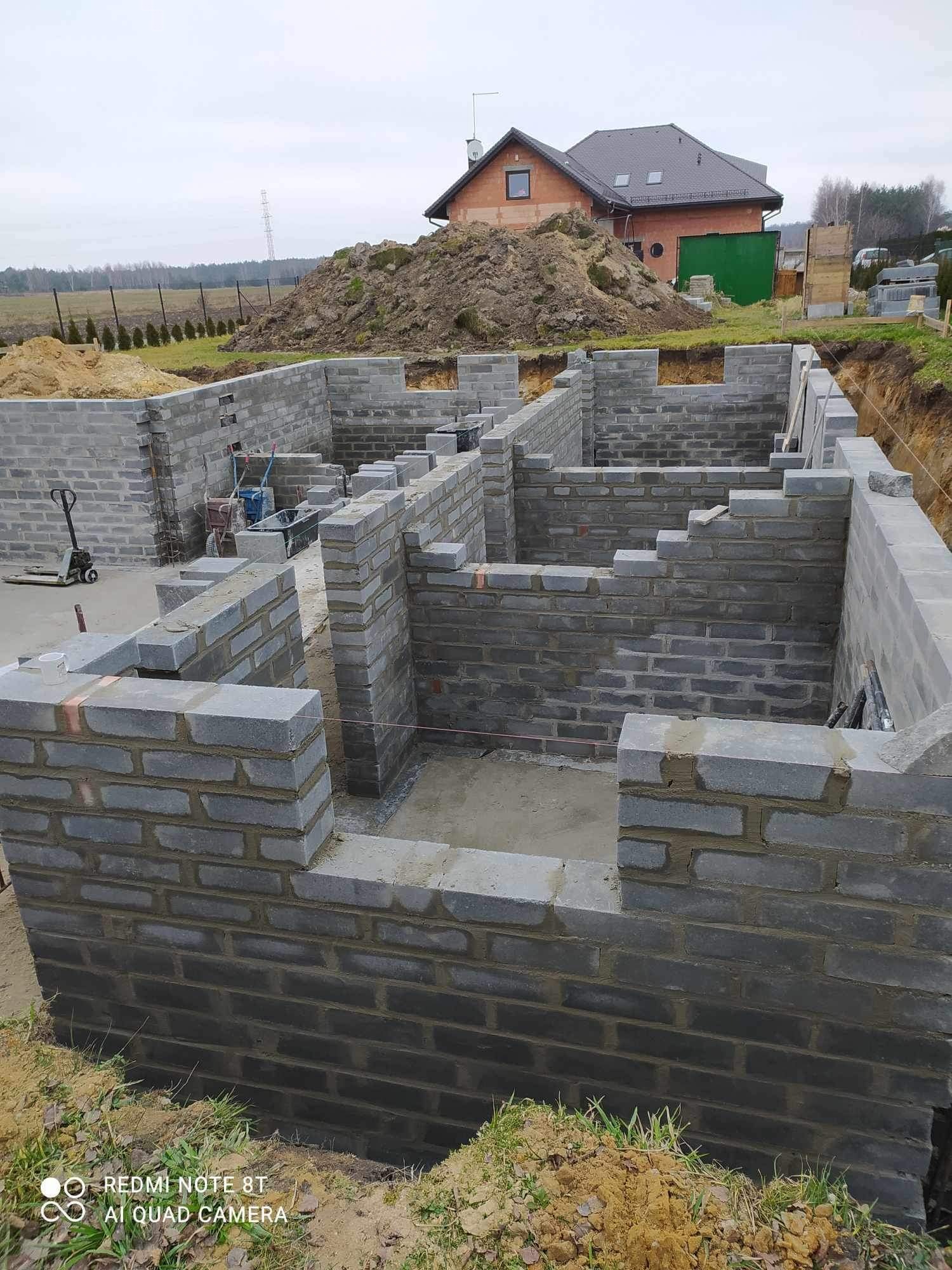Wznoszenie ścian fundamentowych z bloczków betonowych na budowie domu jednorodzinnego; widoczne zbrojenie i poziomowanie, w tle pryzma ziemi i budynek w trakcie budowy.