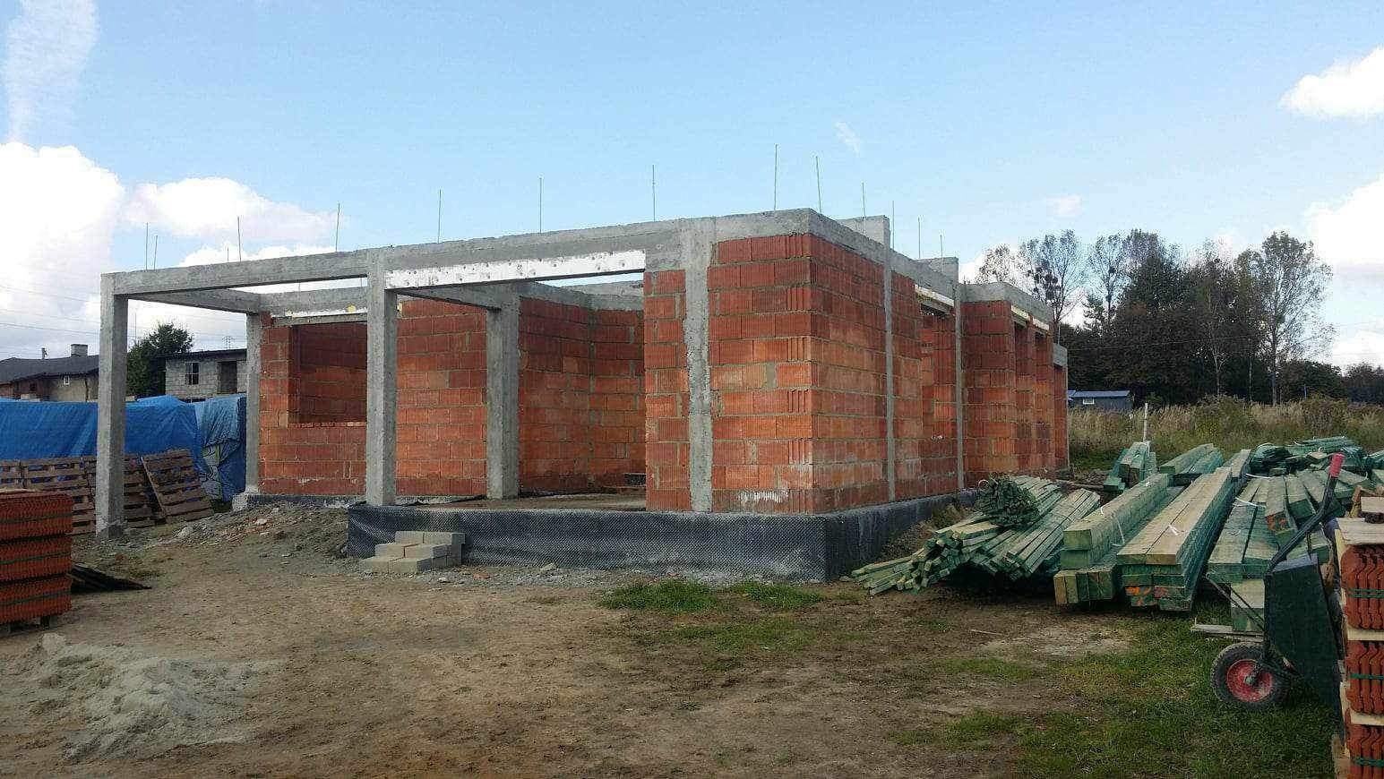 Surowy stan budynku z czerwonej cegły z betonowymi słupami i stropem, widoczne materiały budowlane na zewnątrz, w tle domy i drzewa.