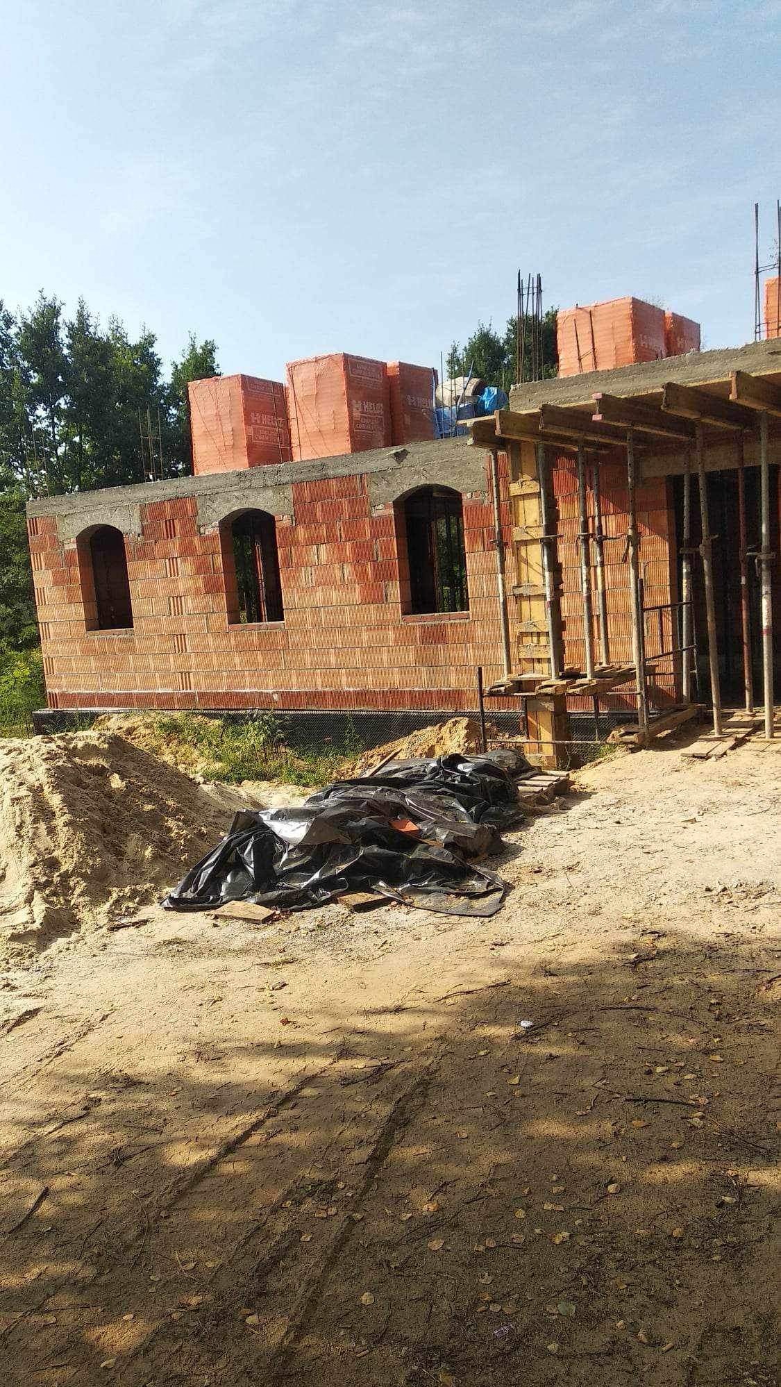 Budowa domu w trakcie wznoszenia ścian z czerwonej cegły, widoczne otwory okienne z łukowym wykończeniem, na górze cegły w pomarańczowych opakowaniach, na ziemi czarna folia i piasek.