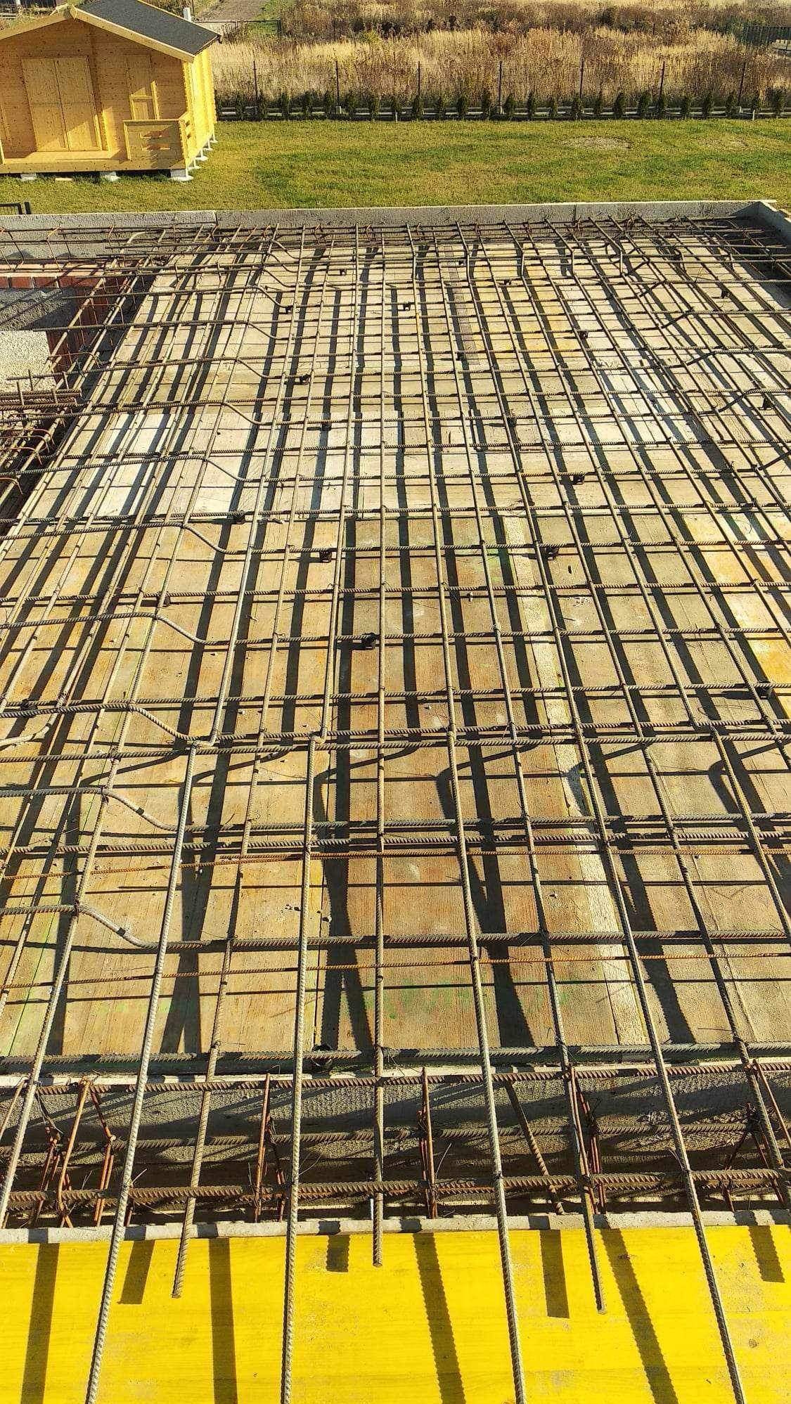 Zbrojenie fundamentów ułożone na żółtych szalunkach, widok z góry na siatkę z prętów stalowych, w tle trawa i budynek gospodarczy.
