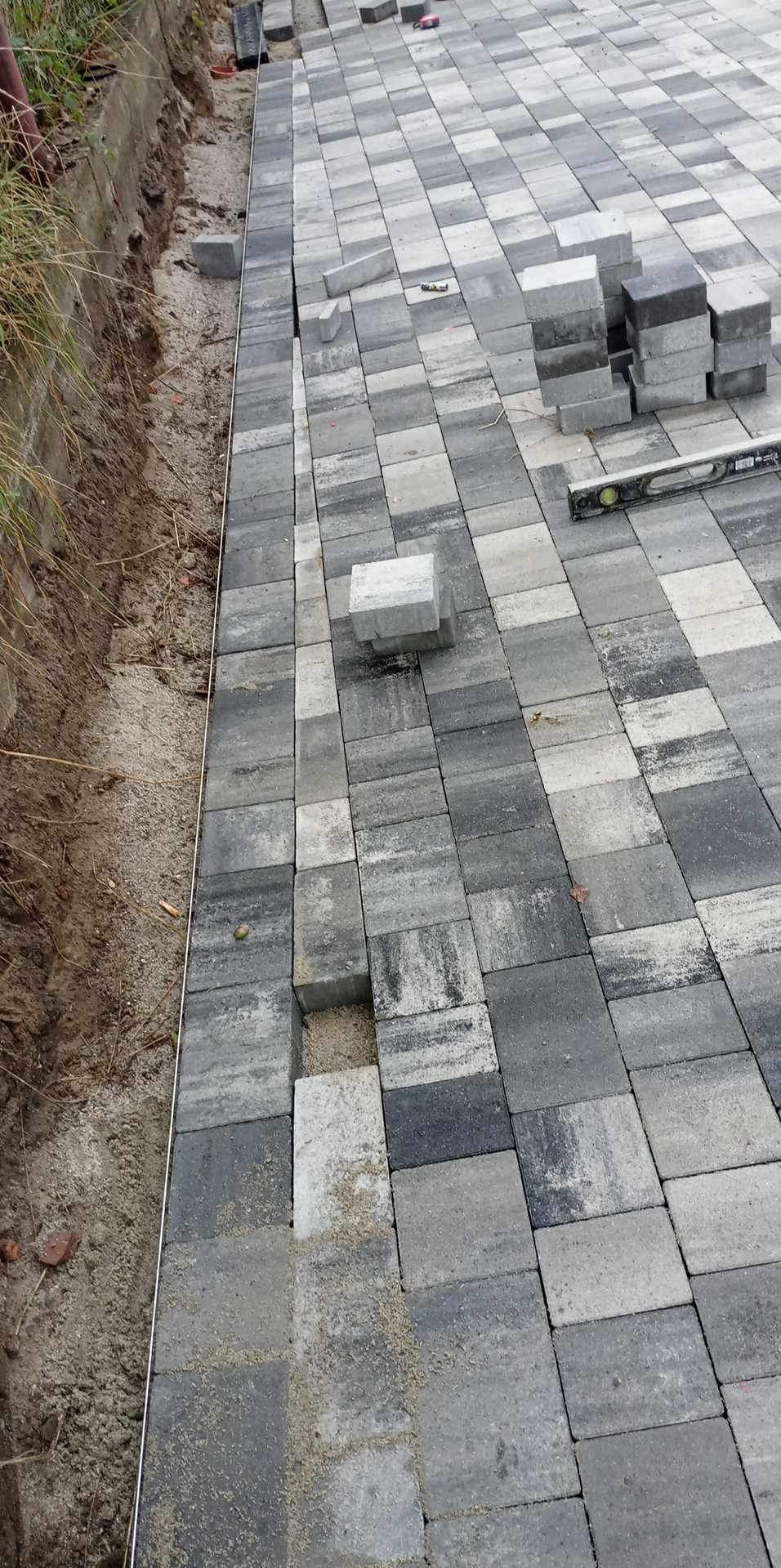 Układanie szarej kostki brukowej na podsypce piaskowej, widoczna poziomica i stos kostki, krawężnik oddzielający od ziemi.