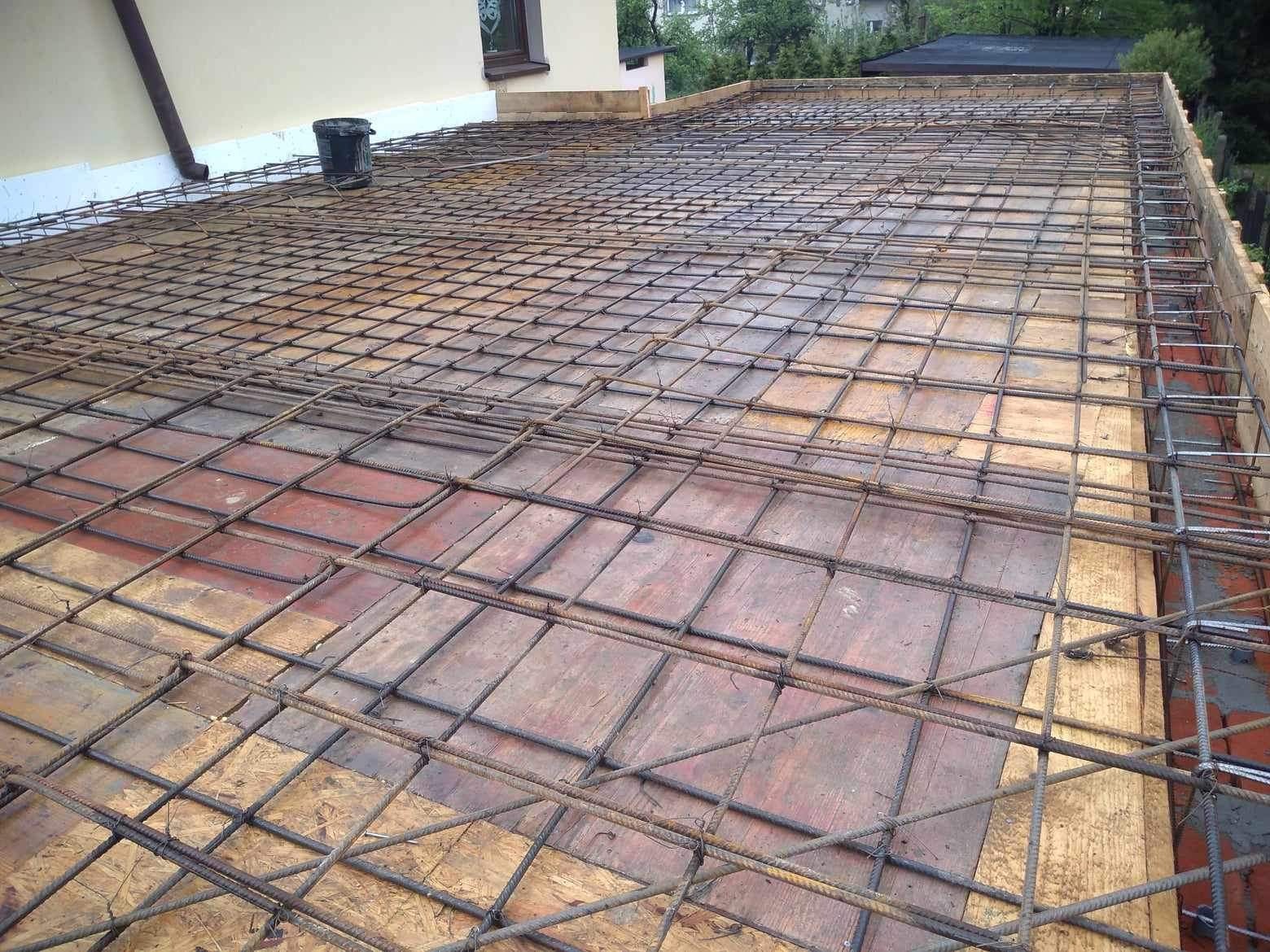 Zbrojenie stropu ułożone na deskowaniu z widocznym obrysem budynku i wiadrem na dachu.