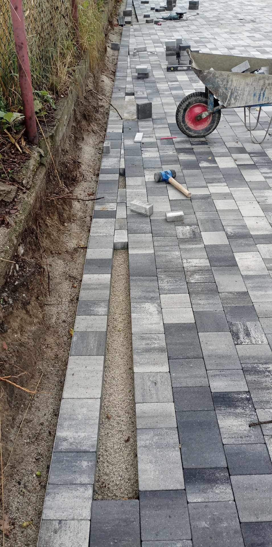 Układanie szarej kostki brukowej na podsypce piaskowej, widoczny gumowy młotek i taczka z czerwoną felgą w tle.