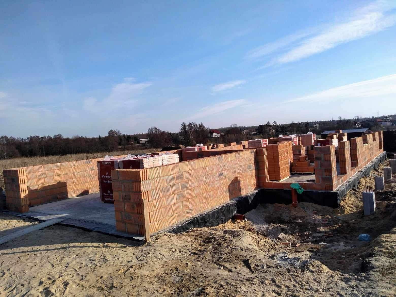 Wznoszone ściany fundamentowe z bloczków ceramicznych Porotherm, zabezpieczone czarną folią hydroizolacyjną, widok na plac budowy w słoneczny dzień.