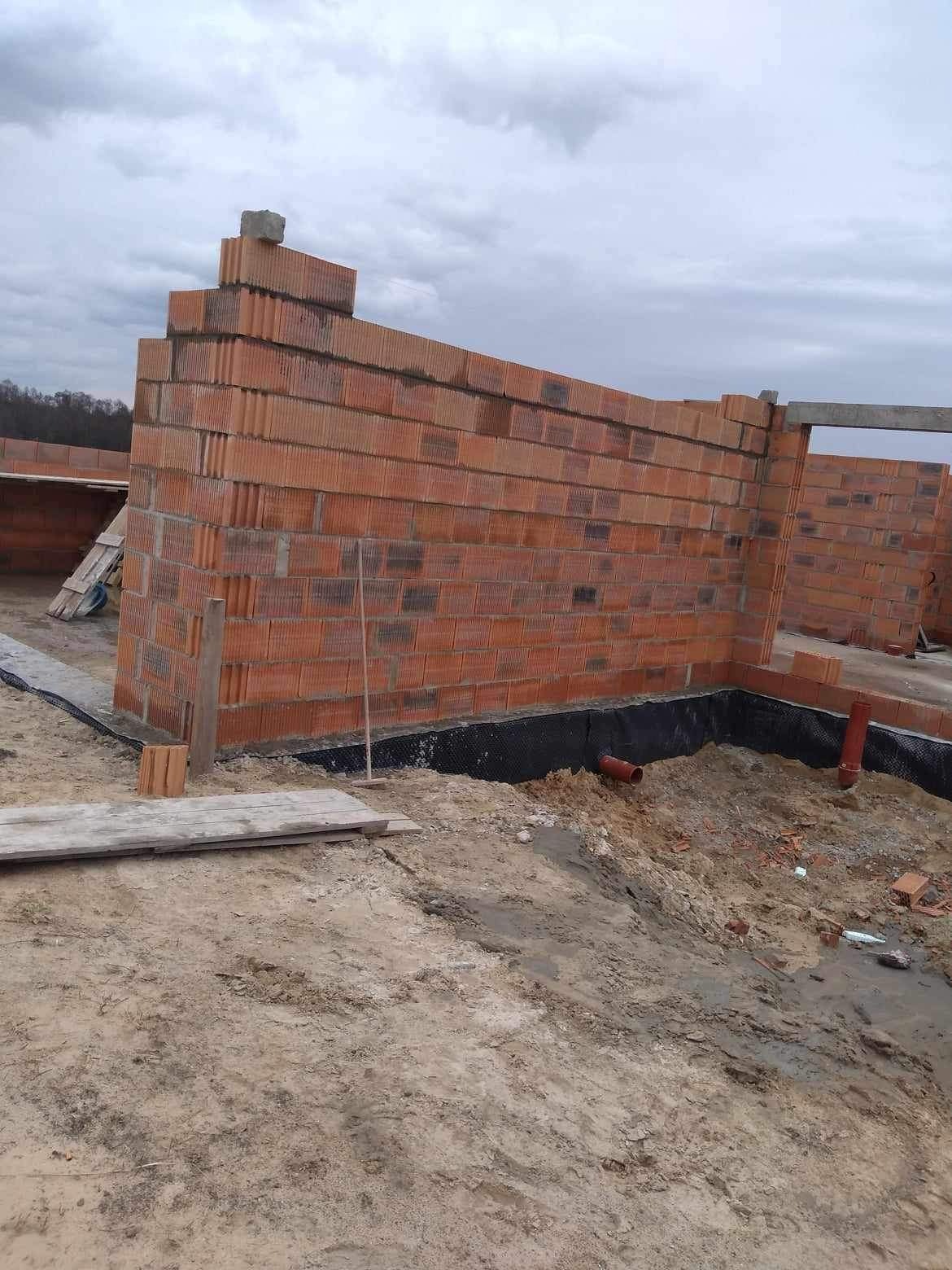 Wznoszone ściany budynku z czerwonej cegły ceramicznej, widoczna izolacja fundamentów w postaci czarnej folii, rury kanalizacyjne i teren budowy w pochmurny dzień.