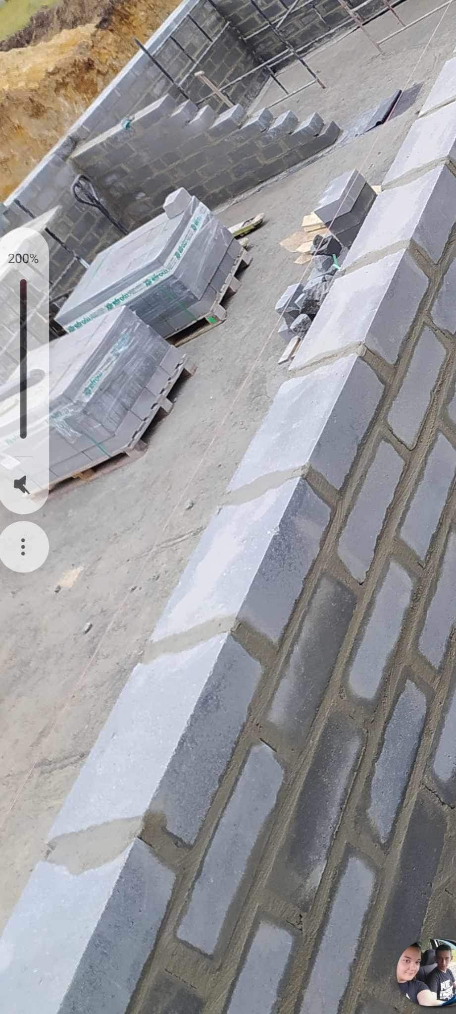 Wznoszenie muru z szarych bloczków betonowych na fundamencie, widoczne palety z zapakowanymi bloczkami w tle