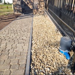 Power Garden - Ułożona kostka brukowa sąsiaduje z pasem wysypanym drobnymi kamykami, przy których zamontowano automat do bramy z niebieską rurą odprowadzającą wodę.