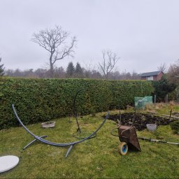 Power Garden - Przygotowanie terenu pod ogród: rozłożony stelaż hamaka, taczka z ziemią, narzędzia ogrodnicze na trawniku przed wysokim żywopłotem i rabatami, pochmurne niebo.