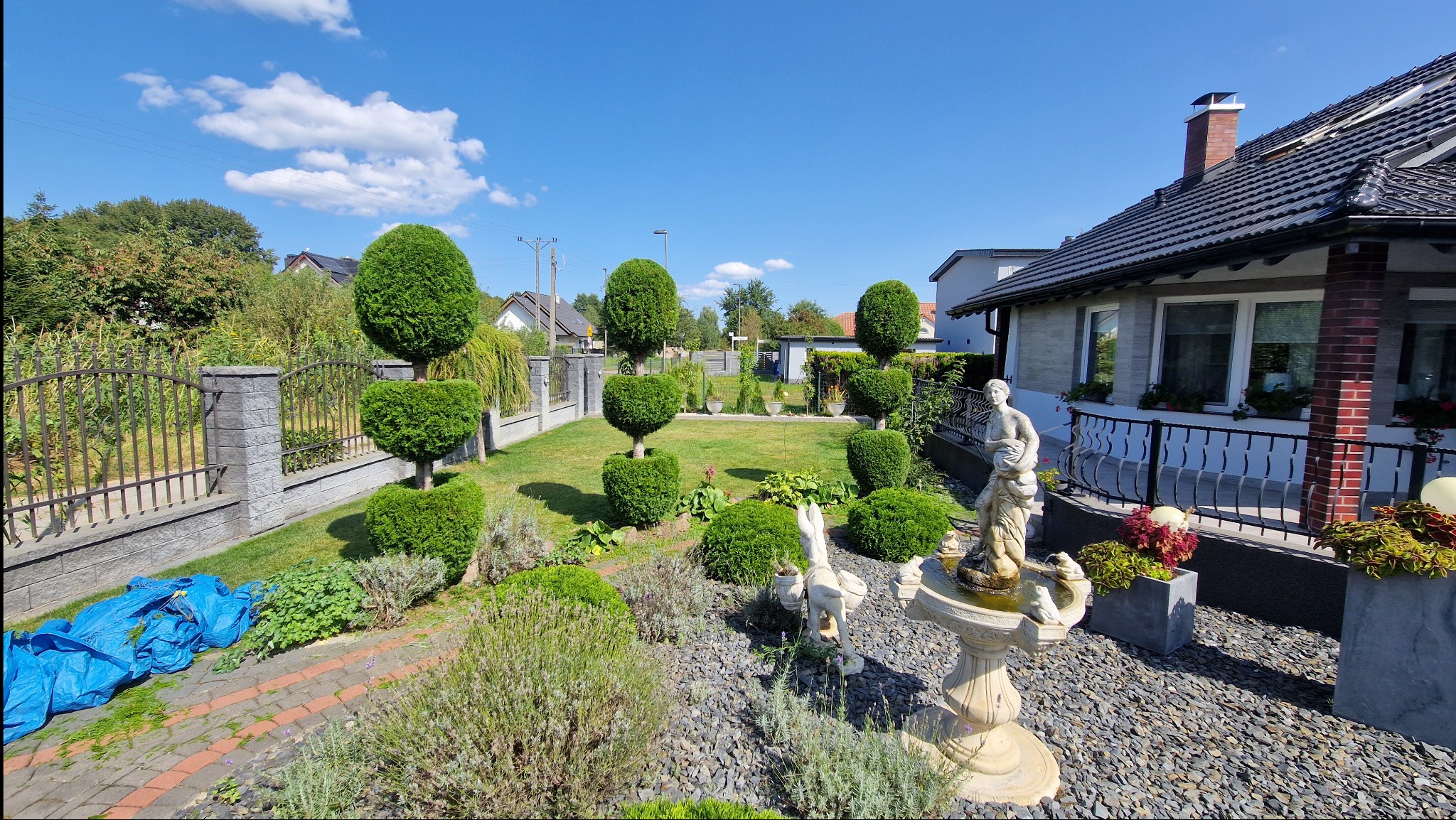 Uformowane krzewy i drzewa w regularnym ogrodzie z fontanną, kamiennym podłożem i zadbanym trawnikiem, widok na dom z czarną dachówką w słoneczny dzień.