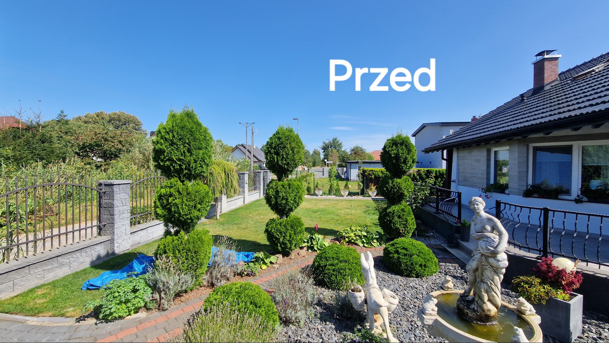 Ogród przydomowy z równo przystrzyżonymi krzewami i drzewami, fontanna z rzeźbą kobiety, trawnik, kostka brukowa i ogrodzenie.