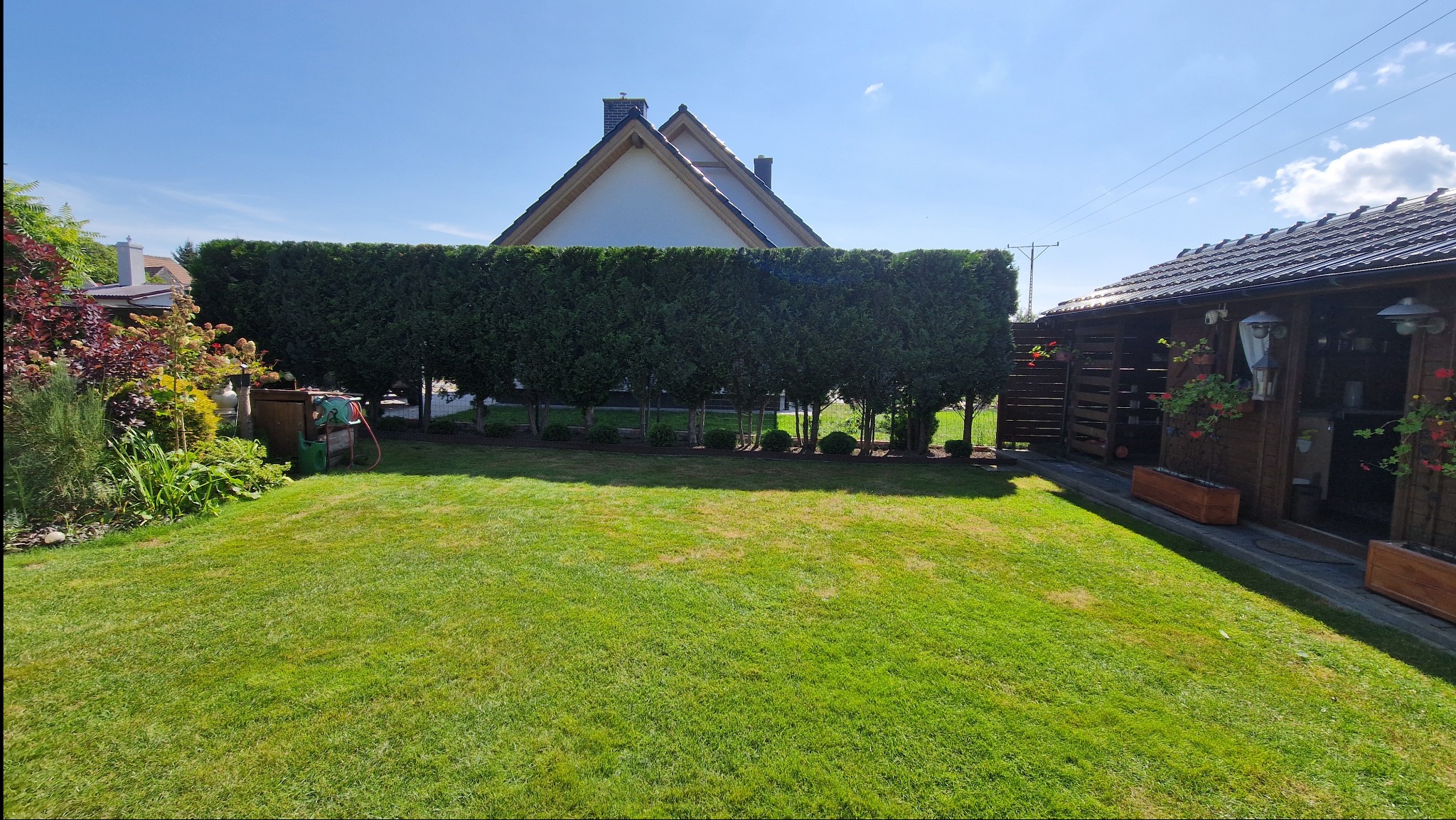 Zadbany ogród z równo przystrzyżonym trawnikiem i żywopłotem z tui, za którym widoczny jest dom. W tle drewniana altana z kwiatami.