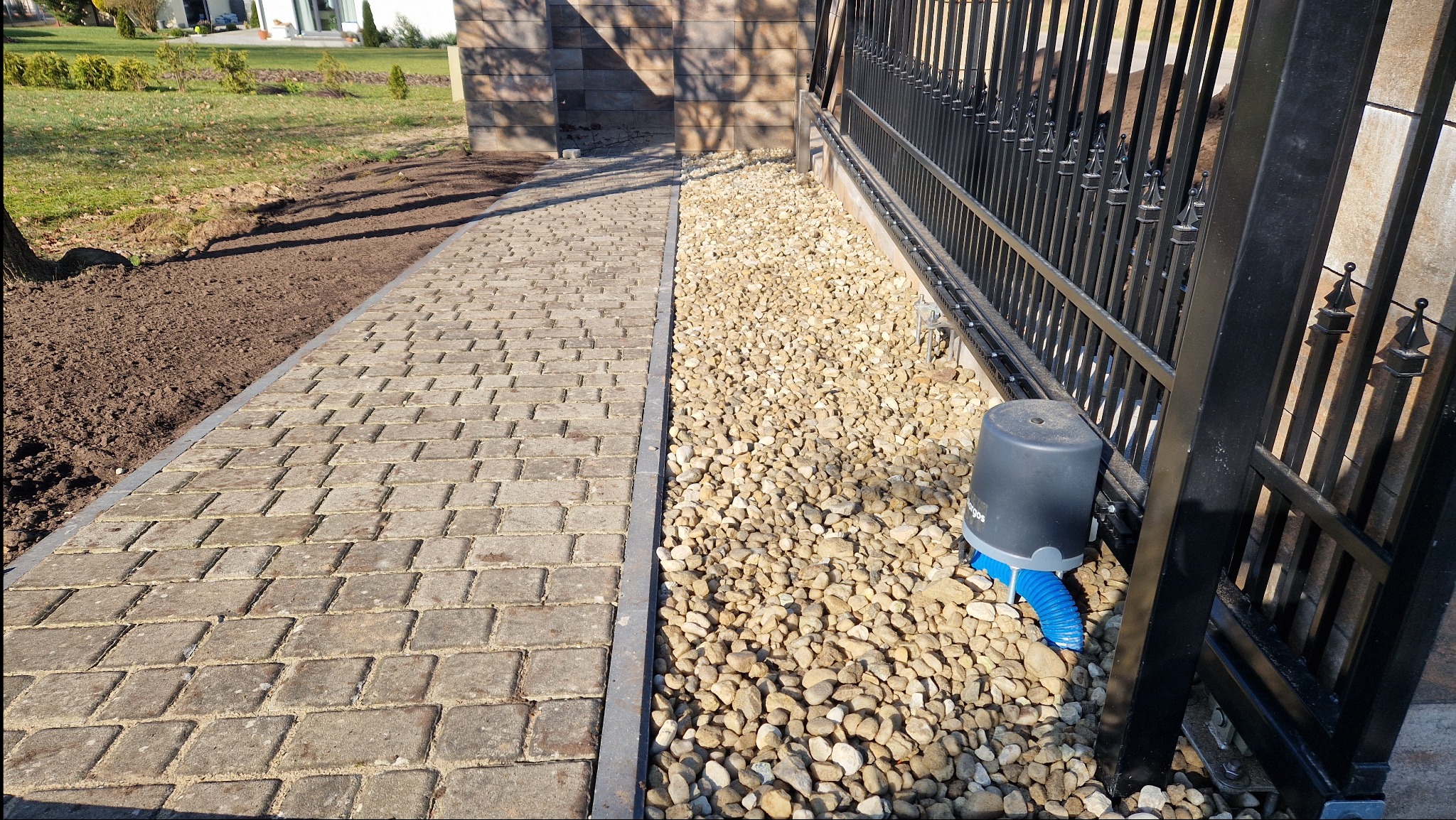 Ułożona kostka brukowa sąsiaduje z pasem wysypanym drobnymi kamykami, przy których zamontowano automat do bramy z niebieską rurą odprowadzającą wodę.