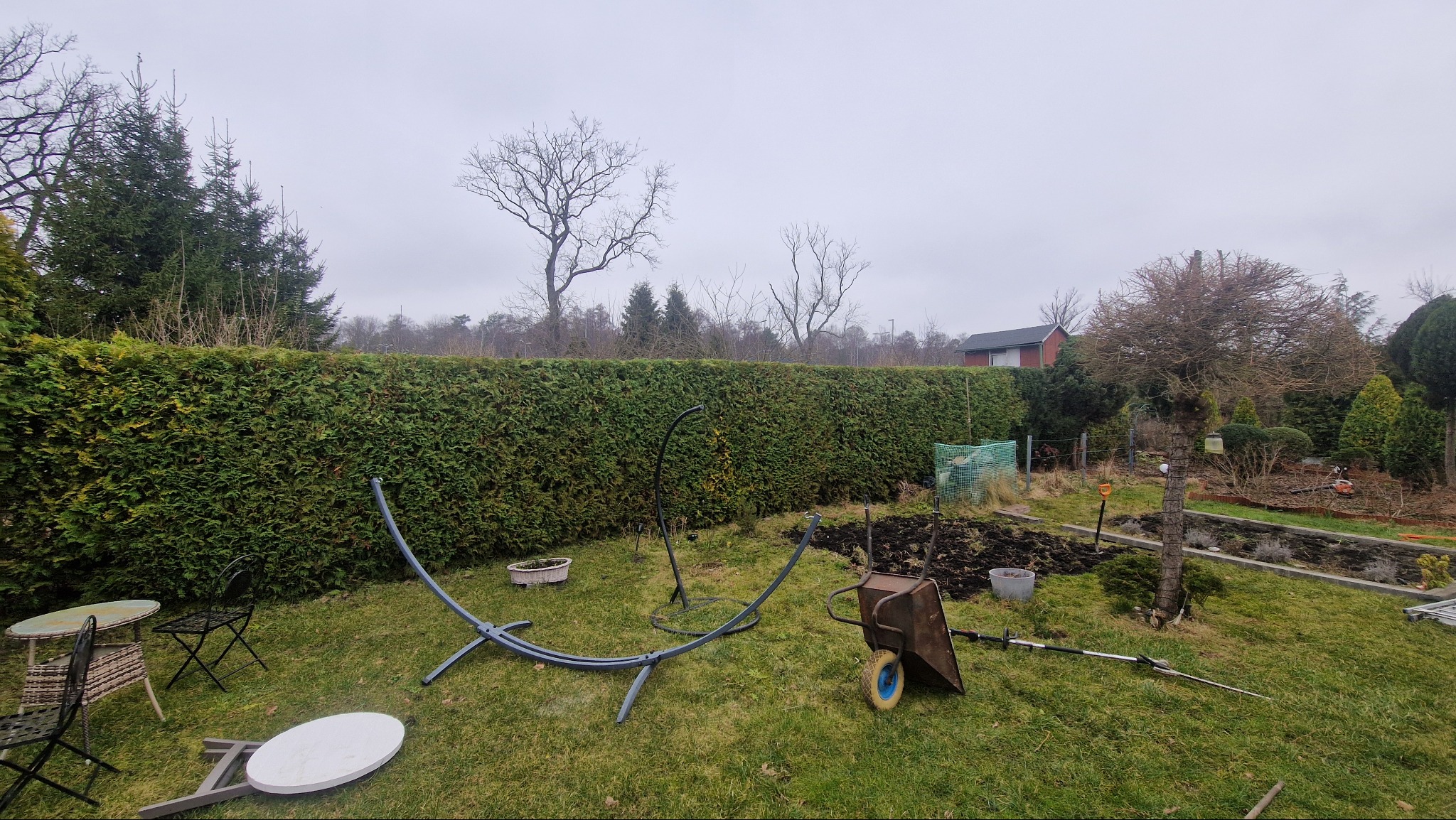 Przygotowanie terenu pod ogród: rozłożony stelaż hamaka, taczka z ziemią, narzędzia ogrodnicze na trawniku przed wysokim żywopłotem i rabatami, pochmurne niebo.