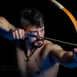 Second Life by Magdalena Baner - Mężczyzna z irokezem i brodą, naciągający łuk, z tatuażem na ramieniu, w złotej bransoletce w kształcie węża, na czarnym tle, z widocznym logo fotografa w lewym dolnym rogu.