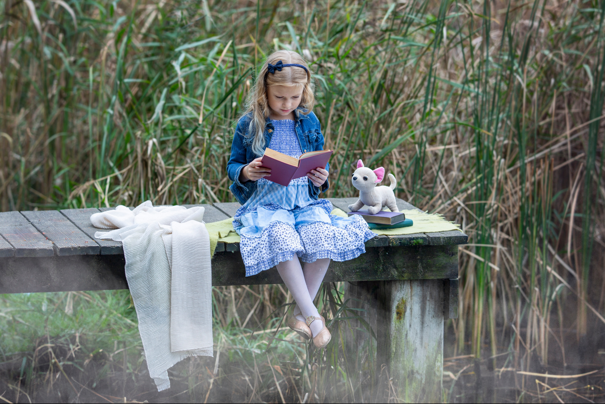 Dziewczynka w dżinsowej kurtce i niebieskiej sukience siedzi na drewnianym pomoście nad wodą, czytając książkę obok pluszowego psa i stosu książek.