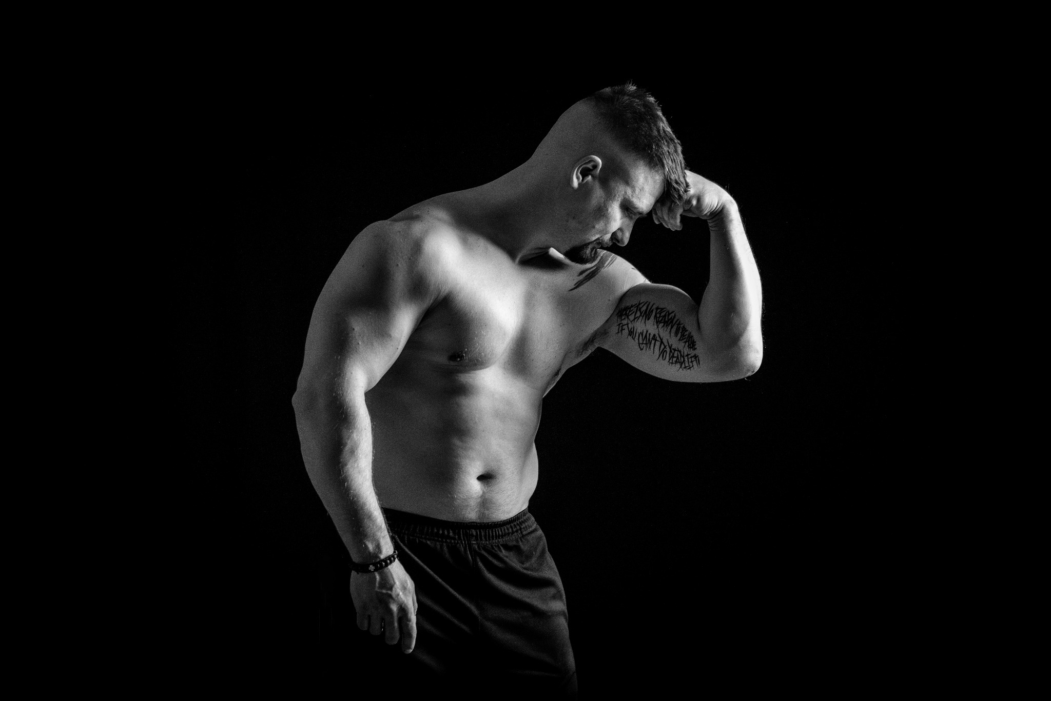 Czarno-białe zdjęcie muskularnego mężczyzny z tatuażem na ramieniu, pozującego na czarnym tle, z głową pochyloną w dół i ręką dotykającą czoła.